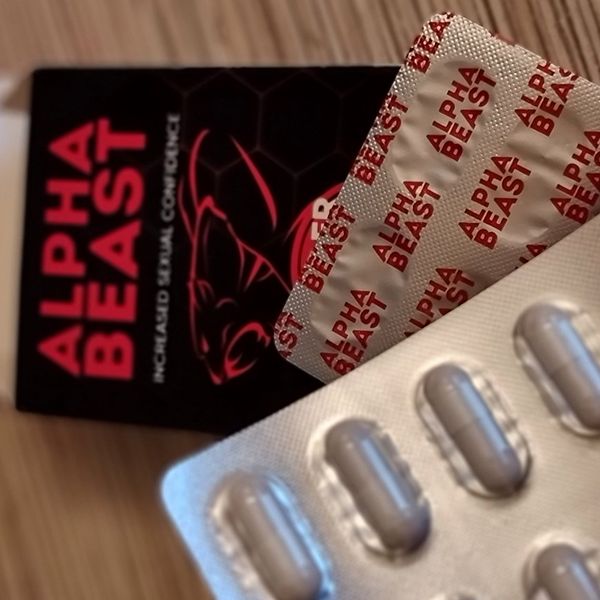Čierna krabička Alpha Beast s červeným nápisom a obrysom šelmy, pred ňou blister s béžovými kapsulami na drevenom stole