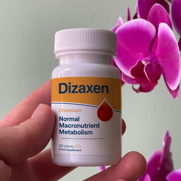 Fľaštička Dizaxen držaná medzi prstami pred sivozelenou stenou, vpravo sýtoružové kvety orchidey