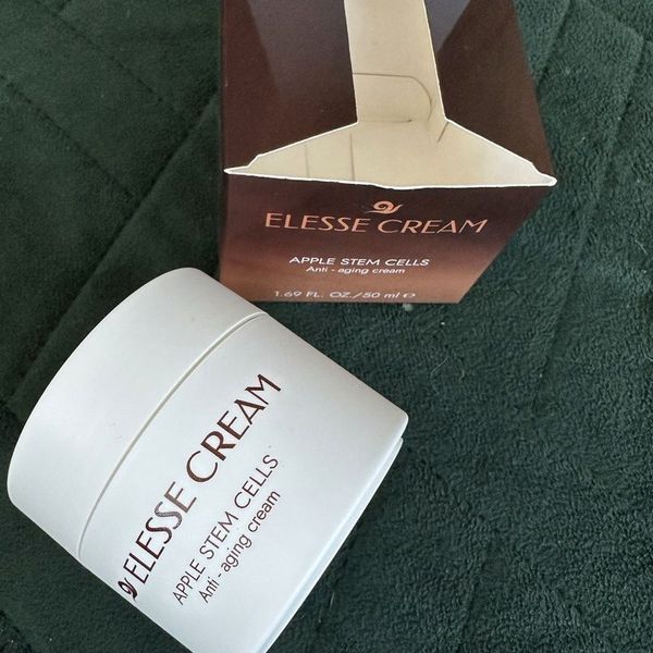Zatvorený biely téglik Elesse Cream ležiaci na boku pri otvorenej hnedej krabičke na tmavozelenej prešívanej deke