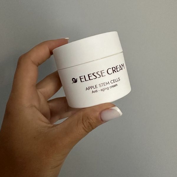 Biely téglik Elesse Cream s bordovým nápisom držaný v prstoch ženskej ruky so svetlými nechtami pred sivou stenou
