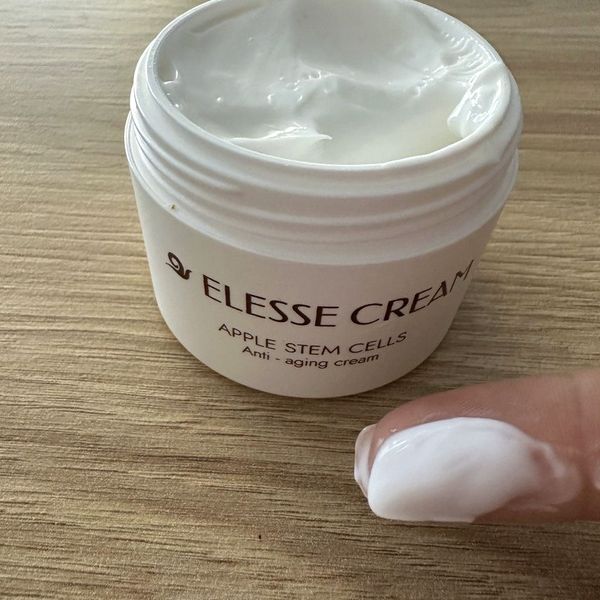 Otvorený téglik Elesse Cream na drevenej doske a kúsok bieleho krému nabraný na prste v popredí