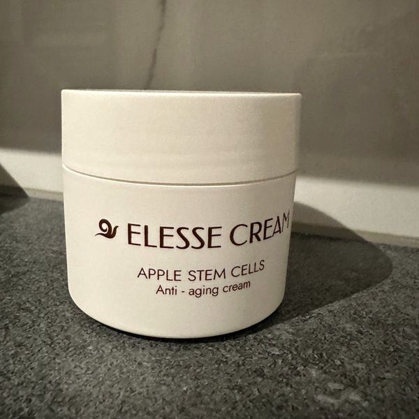 Zatvorený biely téglik Elesse Cream spredu, postavený na tmavosivej kamennej doske pri svetlej stene