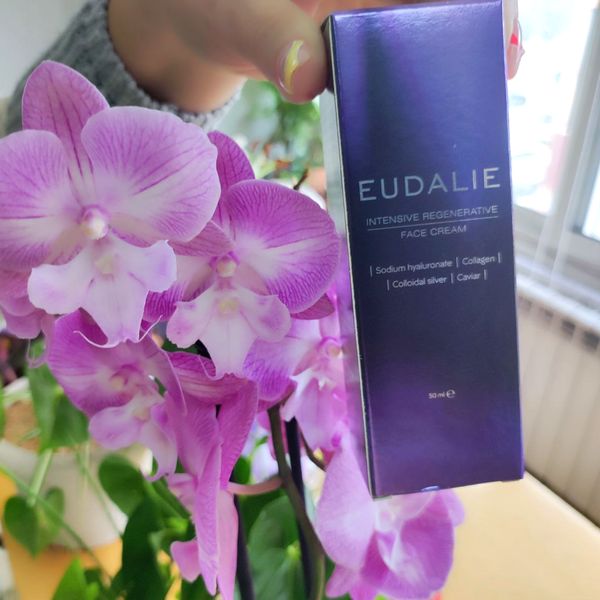 Ruka drží tmavofialovú krabičku krému na tvár Eudalie s údajom 50 ml vedľa kvitnúcej ružovej orchidey