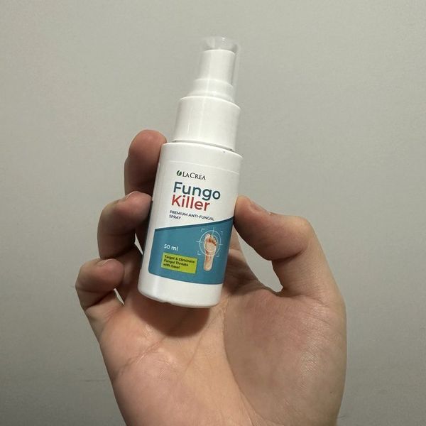 Fľaštička Fungokiller držaná končekmi prstov nad otvorenou dlaňou, etiketa s kresbou chodidla, hladká svetlá stena