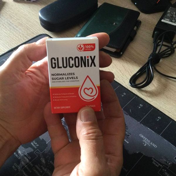 Krabička Gluconix držaná v oboch rukách nad stolom s podložkou s mapou sveta, puzdrom na okuliare a nabíjačkou