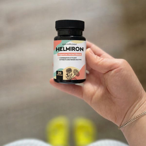 Čierna fľaštička Helmiron s farebnou etiketou a údajom 20 capsules v ženskej dlani, v pozadí rozmazaná podlaha a žlté šľapky