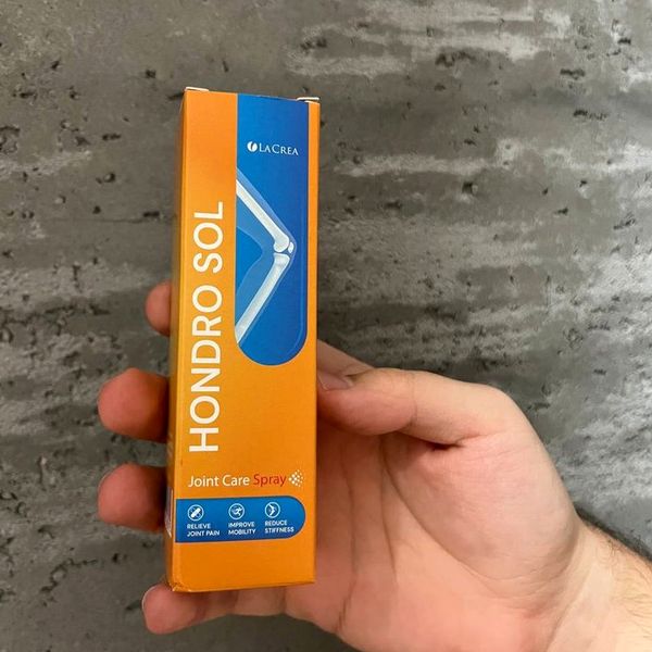 Ruka drží úzku oranžovú krabičku Hondro Sol s modrým pruhom pred sivou betónovou stenou