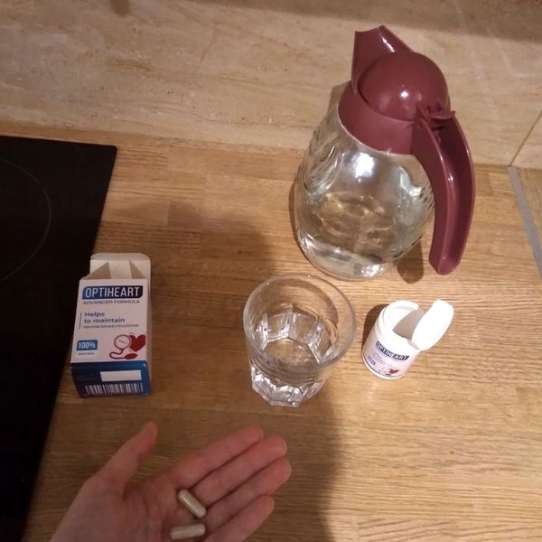 Dve kapsuly na dlani, za nimi krabička a otvorená dóza Optiheart, pohár vody a sklenený džbán na kuchynskej doske