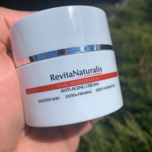 Biely téglik RevitaNaturalis so strieborným pásikom a červeným pruhom na etikete, držaný v dlani, v pozadí zelený ker na slnku