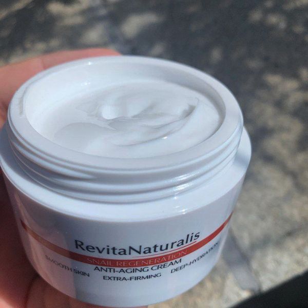 Otvorený téglik RevitaNaturalis plný bieleho krému, držaný v ruke nad slnkom osvetleným chodníkom