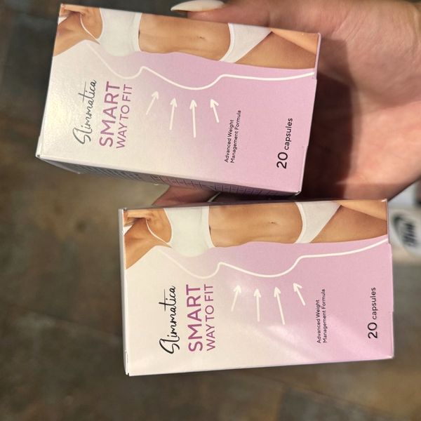 Dve ružovofialové krabičky Slimmatica s údajom 20 capsules, otočené na bok a pridržiavané rukou pred tmavým pozadím