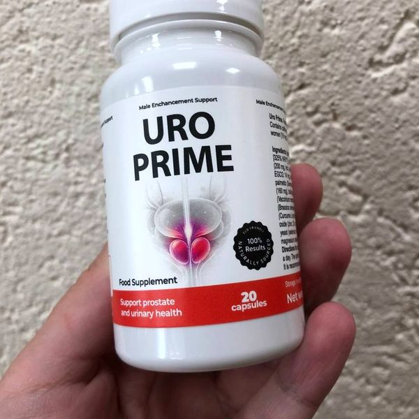Dóza UroPrime držaná v prstoch pred hrubou béžovou omietkou, na boku etikety vidno stĺpec drobného textu