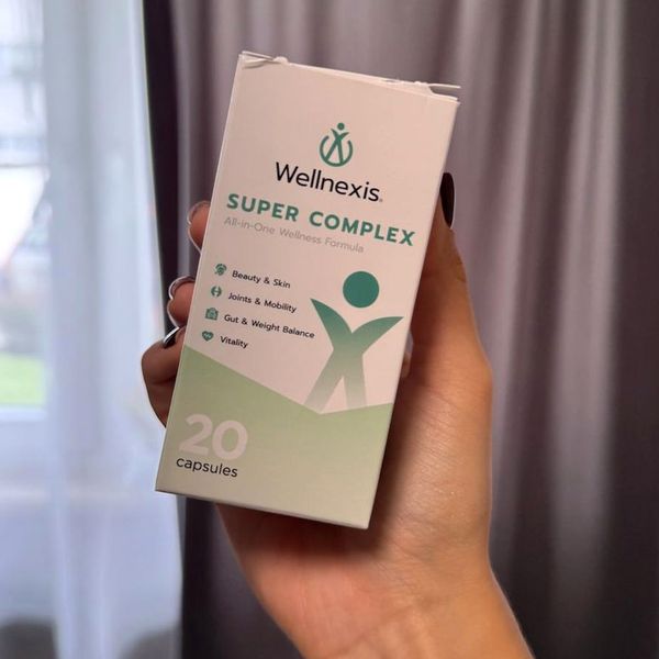 Biela krabička Wellnexis Super Complex so zelenou postavičkou a nápisom 20 capsules v ženskej ruke, v pozadí sivý záves